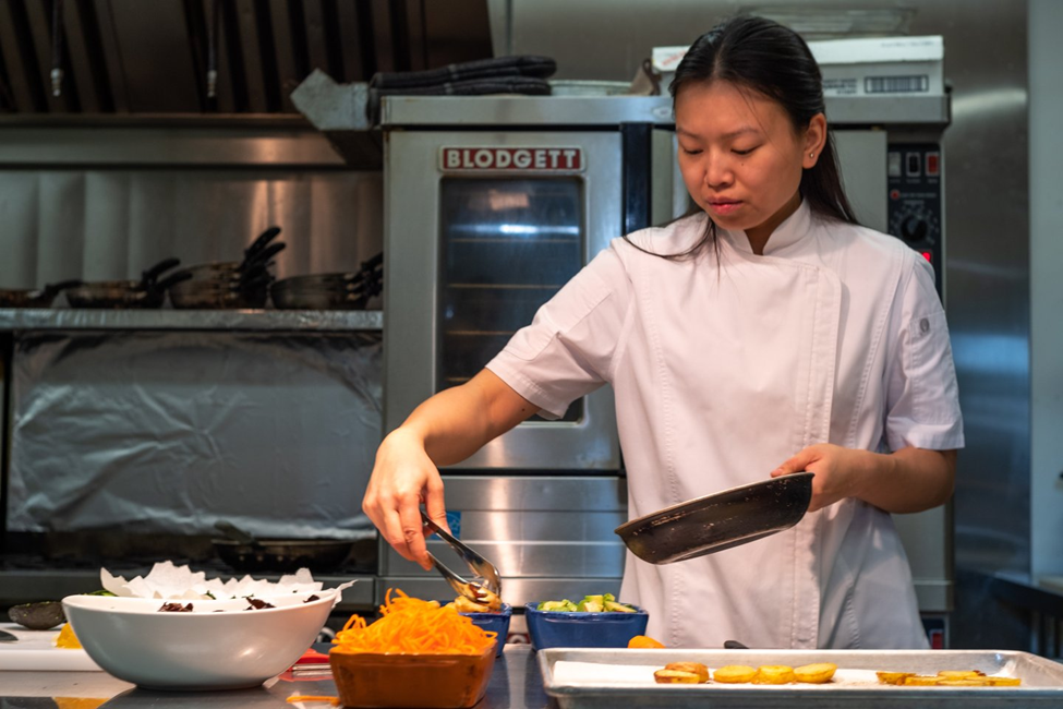 Photograph of a chef in a commercial kitchen preparing food, holding a pan in one hand and using tongs to place ingredients onto a tray. Various bowls with shredded vegetables and other food items are arranged on the counter, with an industrial oven and kitchen equipment visible in the background.
