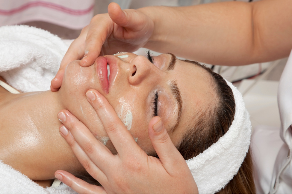 Photograph of a person receiving a facial massage with hands gently pressing on the forehead and cheeks. The scene highlights skincare or relaxation treatment, with a white towel wrapped around the head and a soft, calming environment.