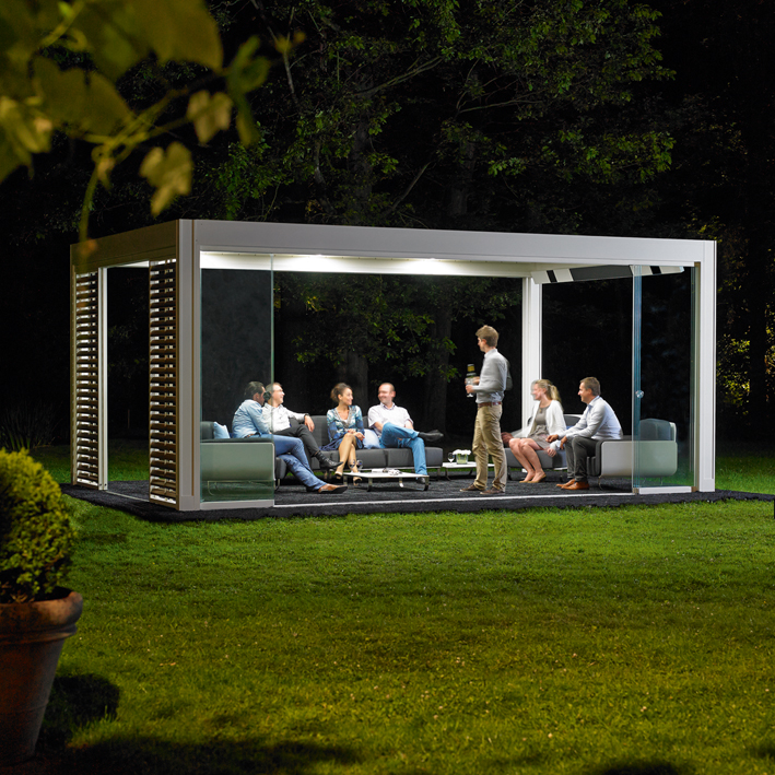 people sitting outside in the nighttime under a bioclimatic pergola in white with lighting and speaker accessories