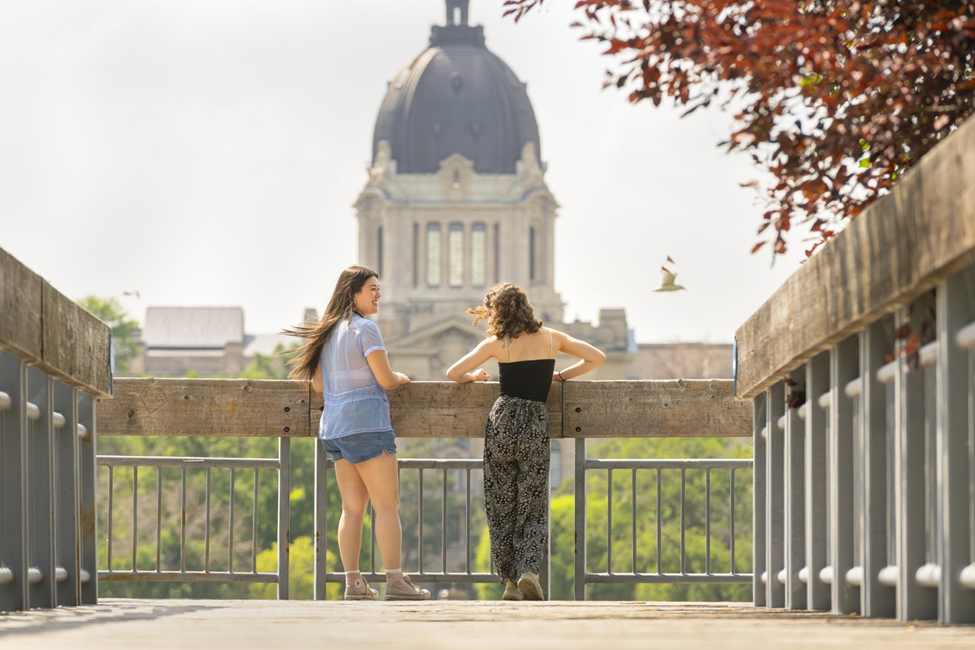 Photograph of two young women standing on a wooden bridge, facing away and looking toward a large domed government building in the background. The scene includes metal railings on both sides of the bridge, green trees, and reddish leaves framing the top right corner, suggesting a peaceful outdoor setting.