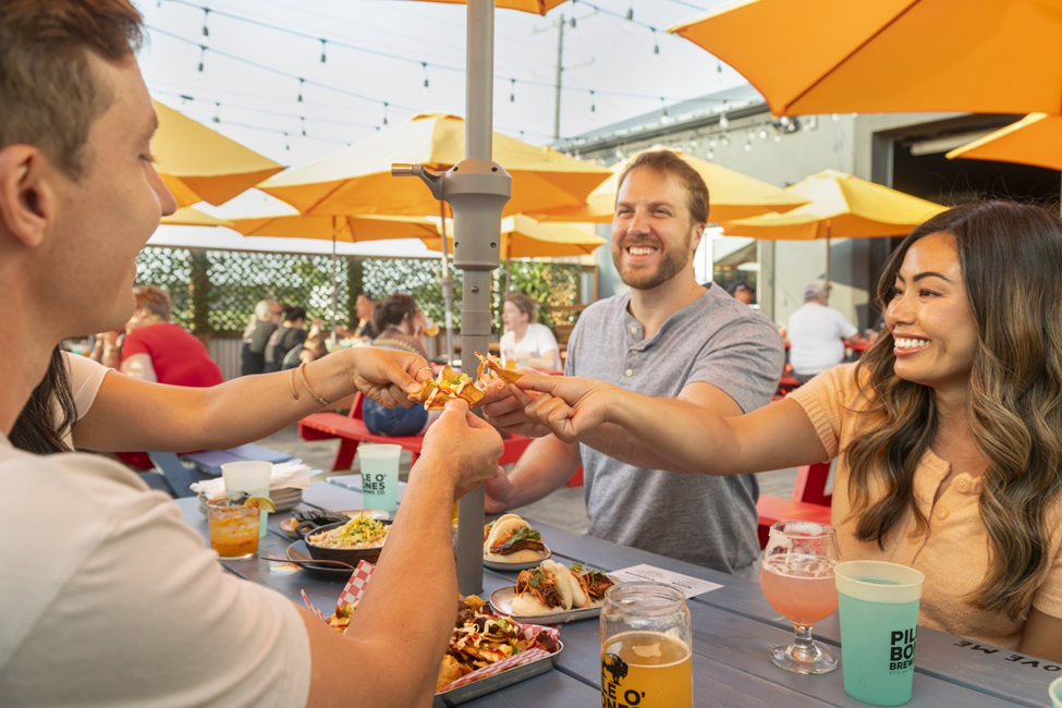 Photograph of a group of people enjoying food and drinks at an outdoor restaurant with orange umbrellas and string lights overhead. The scene highlights social interaction with individuals sharing food, various beverages on the table, and a lively atmosphere in a casual dining setting.