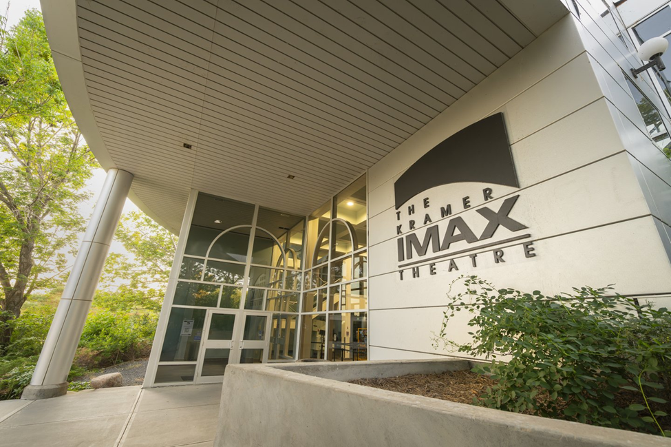 Photograph of an entrance to Kramer IMAX Theatre featuring large glass doors and windows with a white exterior wall displaying bold black signage. Surrounding greenery and a covered walkway with ceiling lights frame the modern architectural design.
